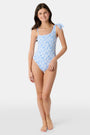 A young girl with long brown hair wears the Roller Rabbit Kids Seaside Knots Jacey One Piece, featuring a blue geometric pattern and a bow on one shoulder, smiling with her hands on her hips against a plain background.