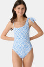 A young girl with long brown hair wears the Roller Rabbit Kids Seaside Knots Jacey One Piece, featuring a blue geometric pattern and a bow on one shoulder, smiling with her hands on her hips against a plain background.