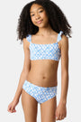 A young girl with curly hair smiles in the Roller Rabbit Kids Seaside Knots Rory Bikini, featuring ruffled shoulder straps and UPF 50+ sun protection, posing against a plain light background.
