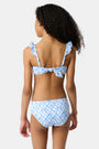 A young girl with curly hair smiles in the Roller Rabbit Kids Seaside Knots Rory Bikini, featuring ruffled shoulder straps and UPF 50+ sun protection, posing against a plain light background.