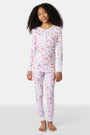 A young girl smiles barefoot in Roller Rabbit Kids Summer Memories Pajamas, made from soft Pima Cotton with playful, colorful prints on white. She stands against a light gray background. Made in Peru.