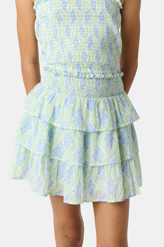 A smiling girl wears the Roller Rabbit Kids Zelda Floral Sami Skirt as she stands by a blue bicycle near a marina, surrounded by others in summer outfits with boats, water, and houses in the background under a clear sky.