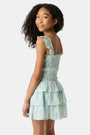 A girl with long curly hair wears the Kids Zelda Floral Tory Top by Roller Rabbit, styled as a light blue and green tiered dress, standing against a plain white background with her hands behind her back.