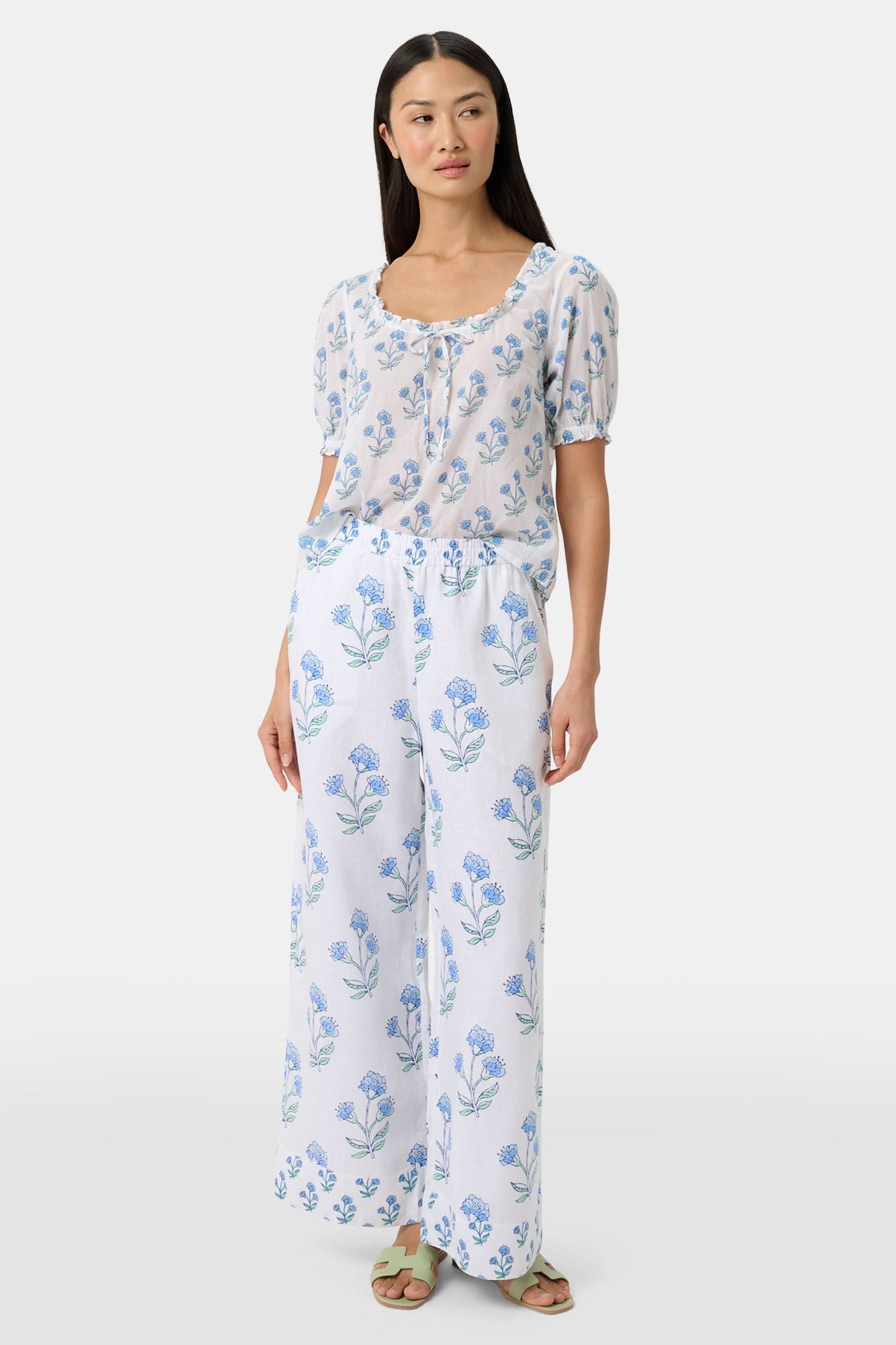A person wears the Roller Rabbit Rosa Floral Kaylee Pant—high-waisted white linen pants with blue floral print and pockets—paired with an off-white top and green sandals, standing against a plain light background.
