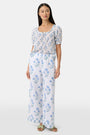 A person wears the Roller Rabbit Rosa Floral Kaylee Pant—high-waisted white linen pants with blue floral print and pockets—paired with an off-white top and green sandals, standing against a plain light background.