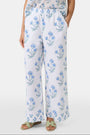 A person wears the Roller Rabbit Rosa Floral Kaylee Pant—high-waisted white linen pants with blue floral print and pockets—paired with an off-white top and green sandals, standing against a plain light background.