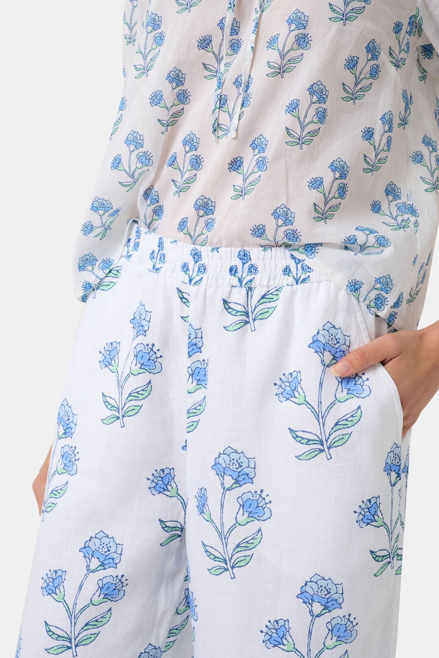 A person wears the Roller Rabbit Rosa Floral Kaylee Pant—high-waisted white linen pants with blue floral print and pockets—paired with an off-white top and green sandals, standing against a plain light background.