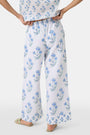 A person wears the Roller Rabbit Rosa Floral Kaylee Pant—high-waisted white linen pants with blue floral print and pockets—paired with an off-white top and green sandals, standing against a plain light background.