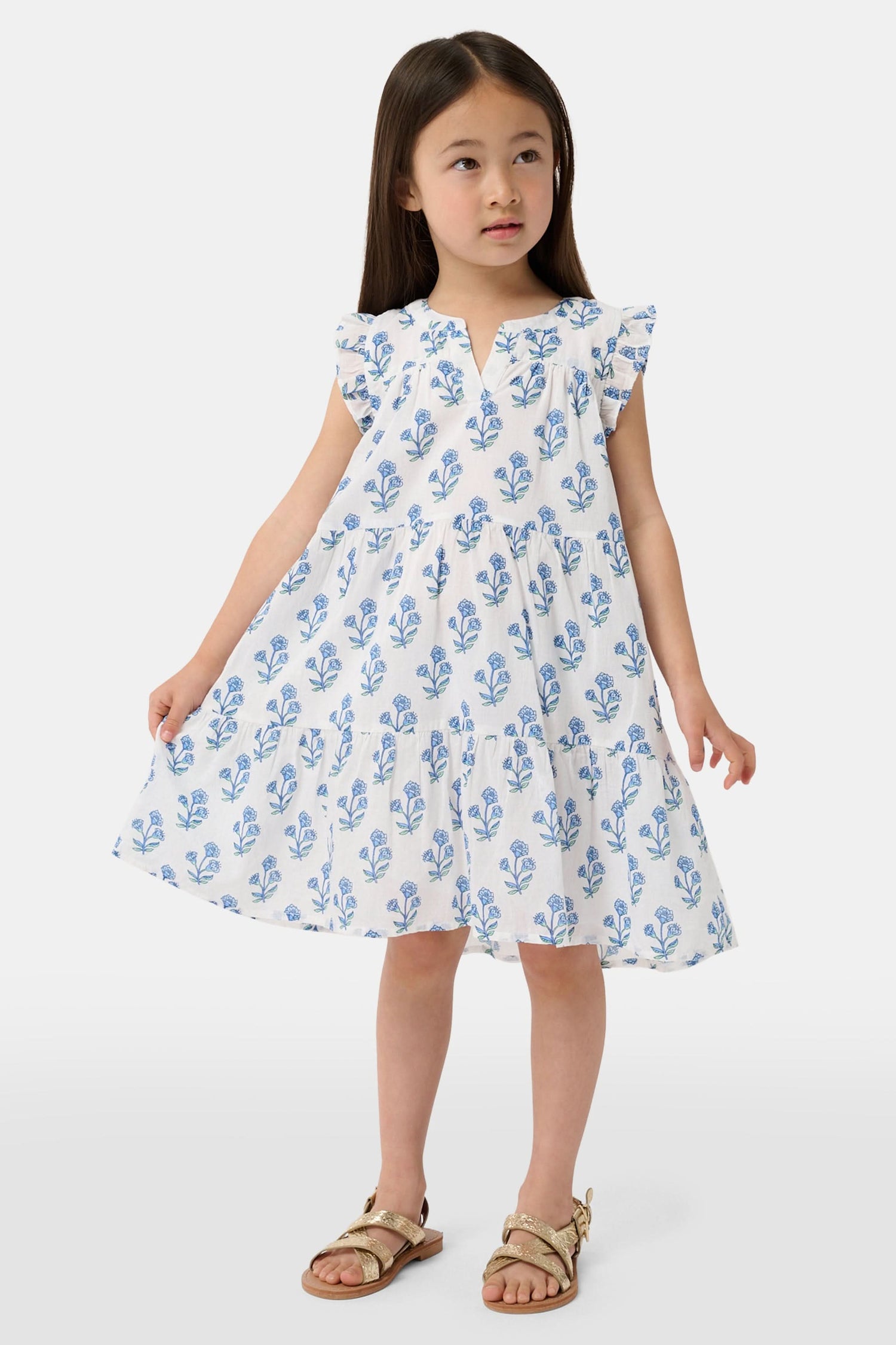 A young girl with long brown hair wears the Roller Rabbit Kids Rosa Ditsy Floral Pami Dress, made from cotton voile with blue-green block print and ruffled sleeves, standing against a plain light background, looking slightly to the side.