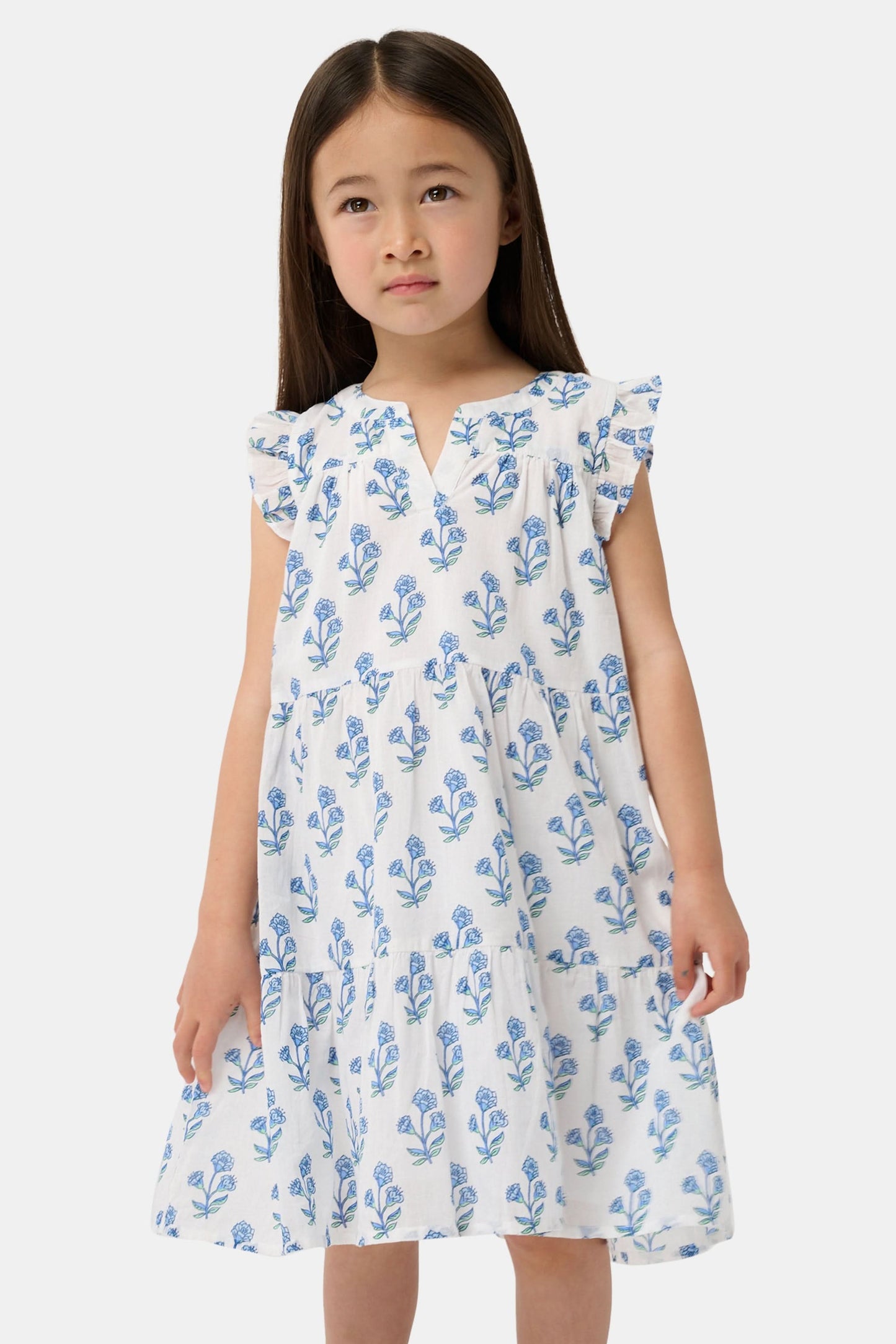 A young girl with long brown hair wears the Roller Rabbit Kids Rosa Ditsy Floral Pami Dress, made from cotton voile with blue-green block print and ruffled sleeves, standing against a plain light background, looking slightly to the side.
