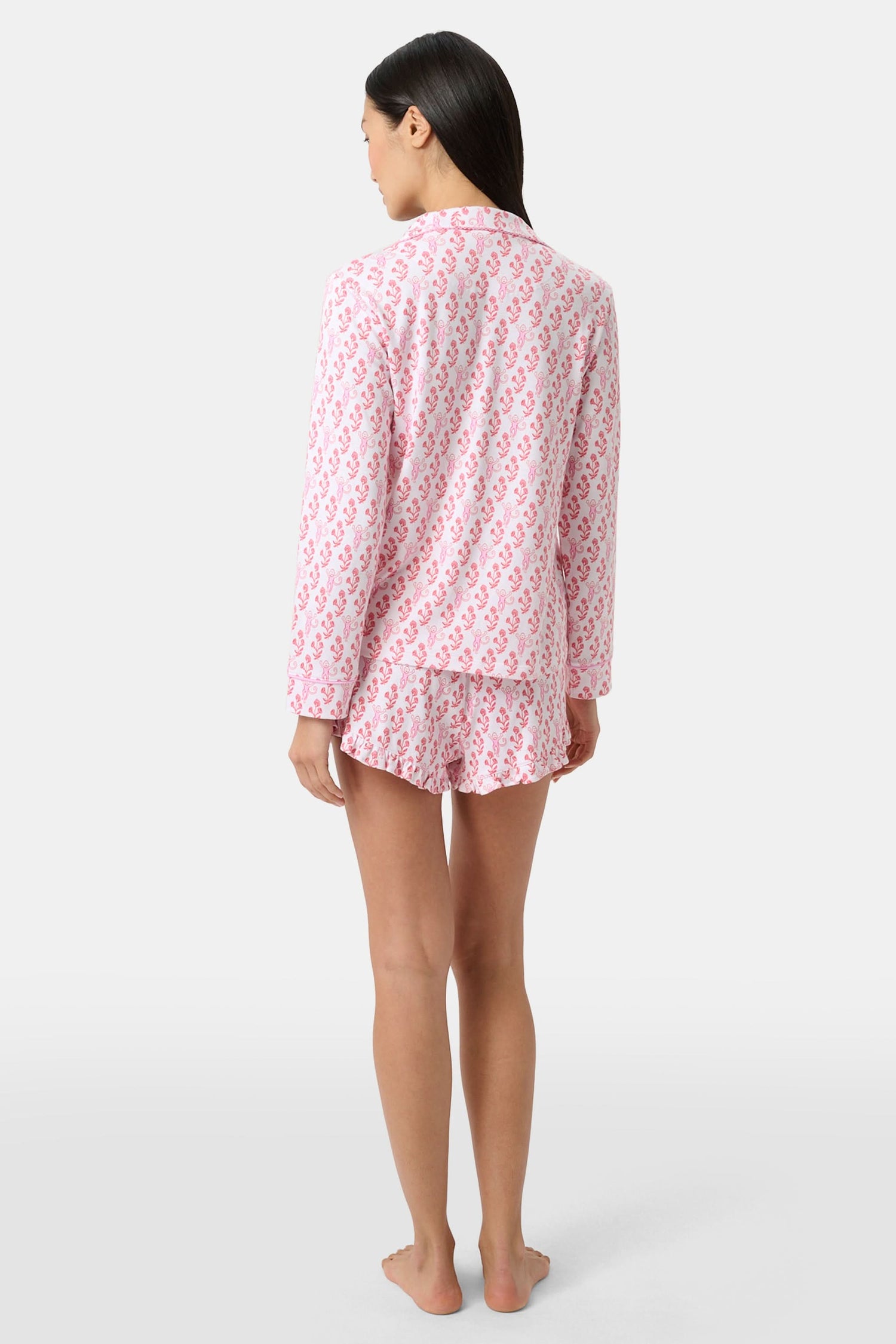 Wearing the Roller Rabbit Rosa Ditsy Monkey Hayden Pajamas—a pink and white set with a small anchor print, long-sleeve button-up top, and ruffled shorts—a person stands against a plain light background, smiling slightly.