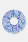 The Rosa Ditsy Monkey Scrunchie by Roller Rabbit is light blue with a playful pink monkey and floral pattern. This soft, gentle scrunchie comes in a circular shape and is displayed against a white background.