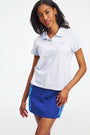 Two women stand on a tennis court wearing Roller Rabbit Pique Polo Shirts; one pairs hers with a blue patterned skirt and navy scarf, the other with a white skirt. A building appears in the background.