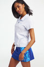 Two women stand on a tennis court wearing Roller Rabbit Pique Polo Shirts; one pairs hers with a blue patterned skirt and navy scarf, the other with a white skirt. A building appears in the background.