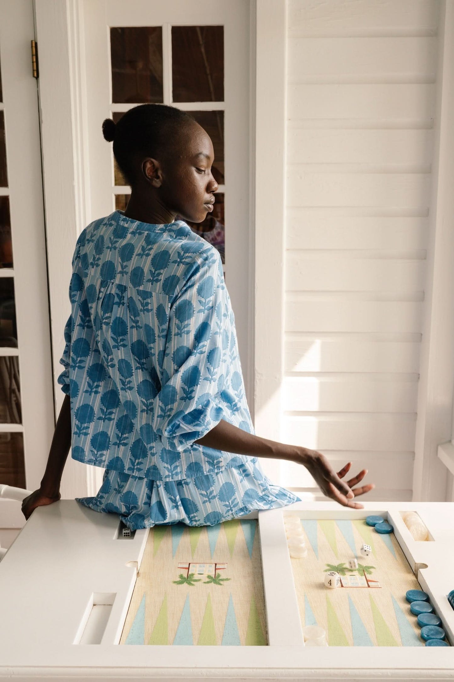 A woman models the Linna Floral Jasper Top by Roller Rabbit, a blue long-sleeve blouse, paired with white shorts and poses against a plain white background, smiling gently at the camera.