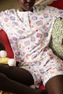Wearing the Tinned or Twist Ashley Top, a white Pima-modal henley pajama shirt with colorful food and drink illustrations, a person sits on a couch with patterned cushions.