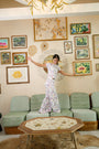 A woman in ultra-soft Tinned or Twist Boden Pants balances on a green sofa in a cozy living room with colorful framed art on the walls and a rattan coffee table in front.