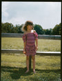 A young child wears the Roller Rabbit Kids Marquis Floral Bibi Skirt paired with maroon heart-accented shoes, standing against a plain white background.