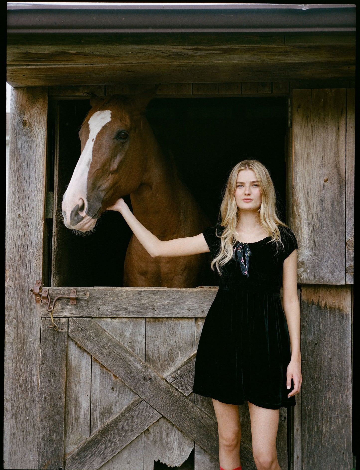 A woman faces forward in the Roller Rabbit Velvet Tilly Dress, a short-sleeve black velvet mini with a floral bow at the neckline, paired with black platform sandals, set against a plain light background.