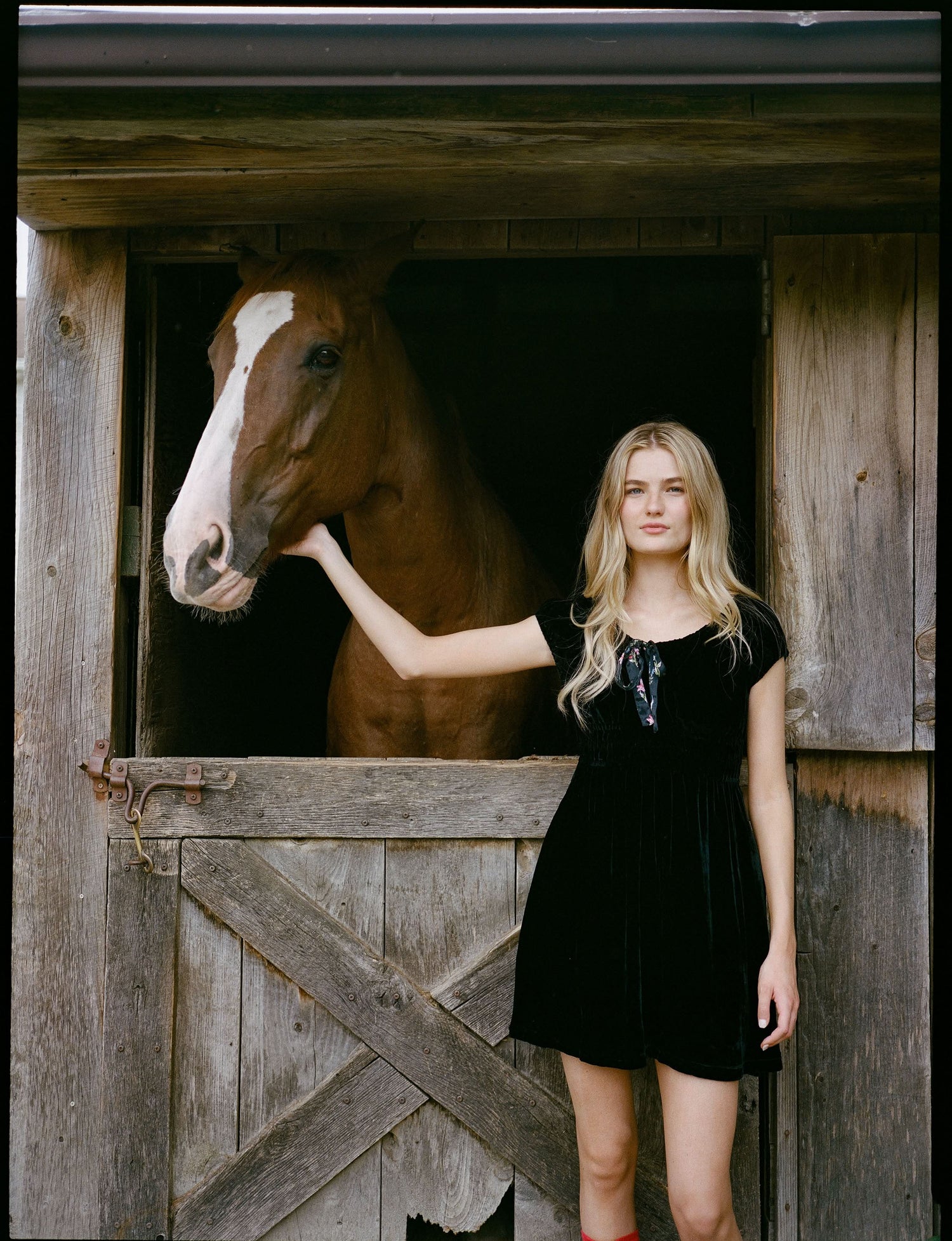A woman faces forward in the Roller Rabbit Velvet Tilly Dress, a short-sleeve black velvet mini with a floral bow at the neckline, paired with black platform sandals, set against a plain light background.