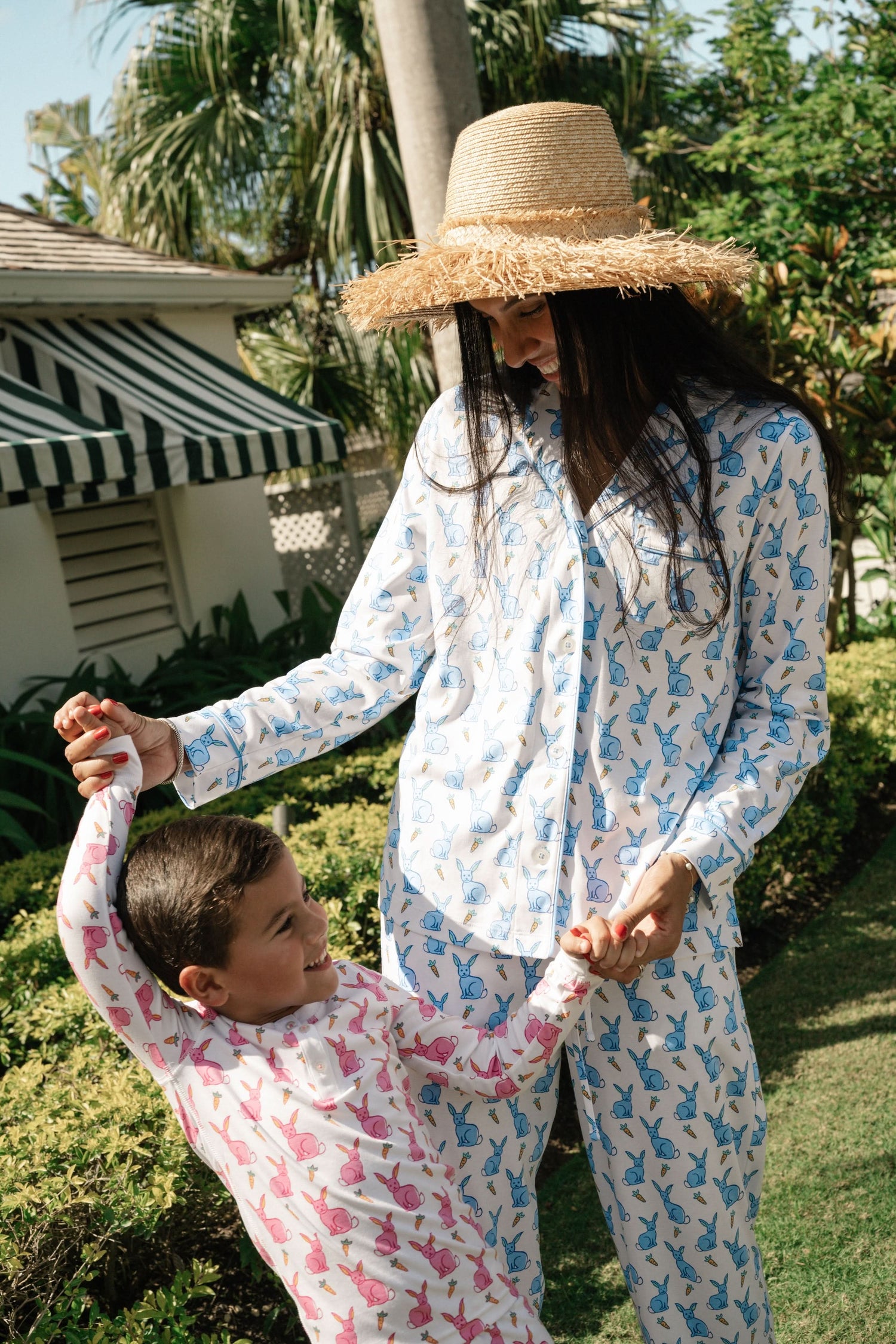 A woman with long brown hair stands barefoot in the Roller Rabbit Romy the Rabbit Long Sleeve Polo Pajamas—a matching white set with a blue bunny print, featuring a button-up top and loose-fitting pants. She has a neutral expression.