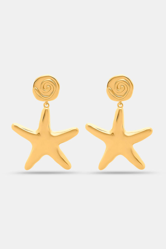 Close-up of a person’s ear with Roller Rabbit Starfish Drop Earrings—gold-tone danglers featuring a small rose detail—against curly dark hair and tan skin.