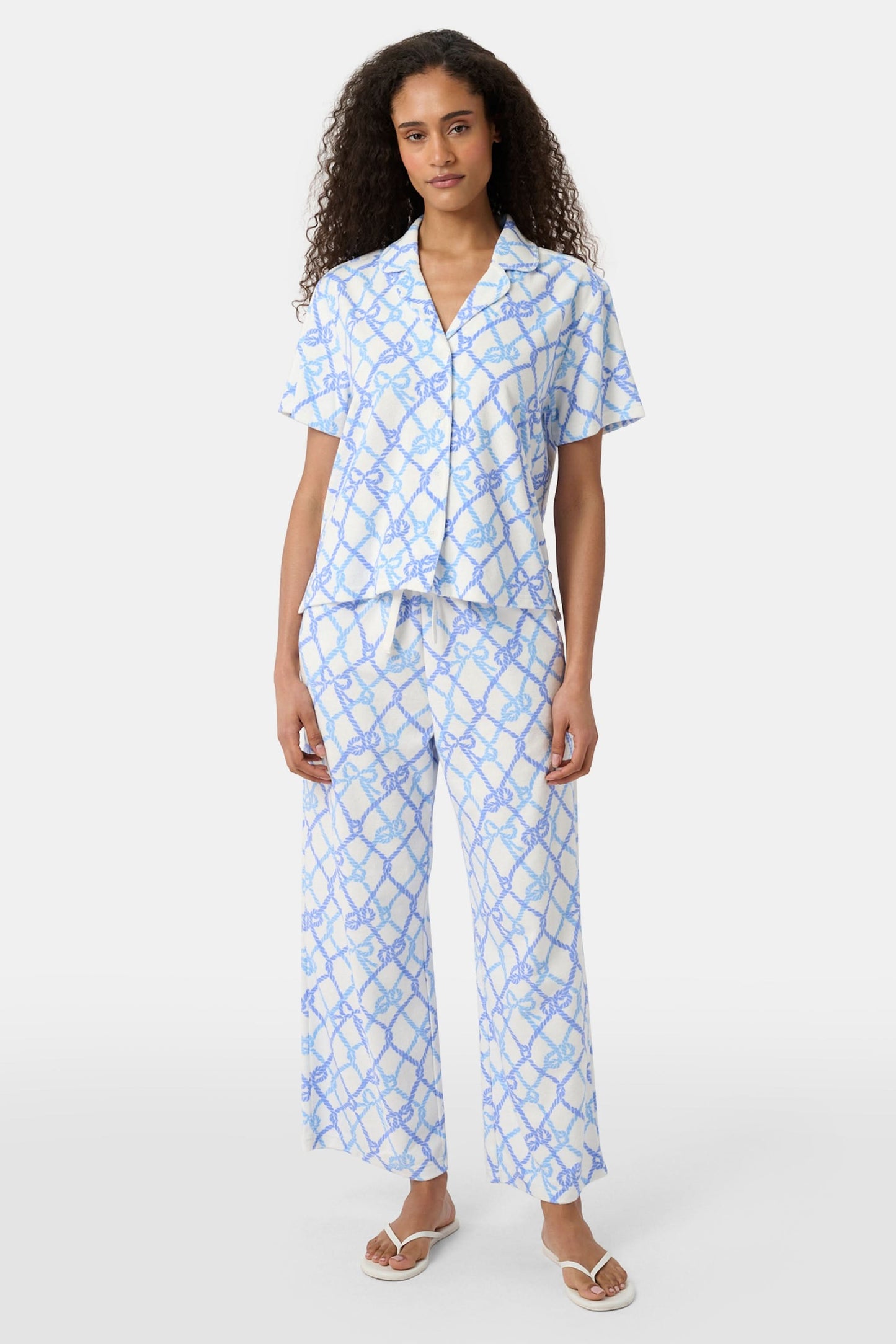 A person wears the Roller Rabbit Seaside Knots Mara Pant, featuring a tie waist and wide legs in loose white fabric with a light blue geometric chain pattern, paired with a matching top and white sandals against a plain white background.