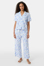 A person wears the Roller Rabbit Seaside Knots Mara Pant, featuring a tie waist and wide legs in loose white fabric with a light blue geometric chain pattern, paired with a matching top and white sandals against a plain white background.