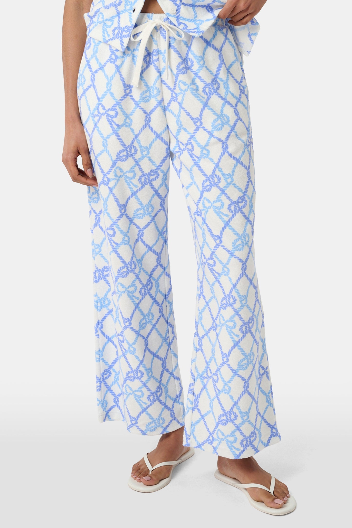 A person wears the Roller Rabbit Seaside Knots Mara Pant, featuring a tie waist and wide legs in loose white fabric with a light blue geometric chain pattern, paired with a matching top and white sandals against a plain white background.