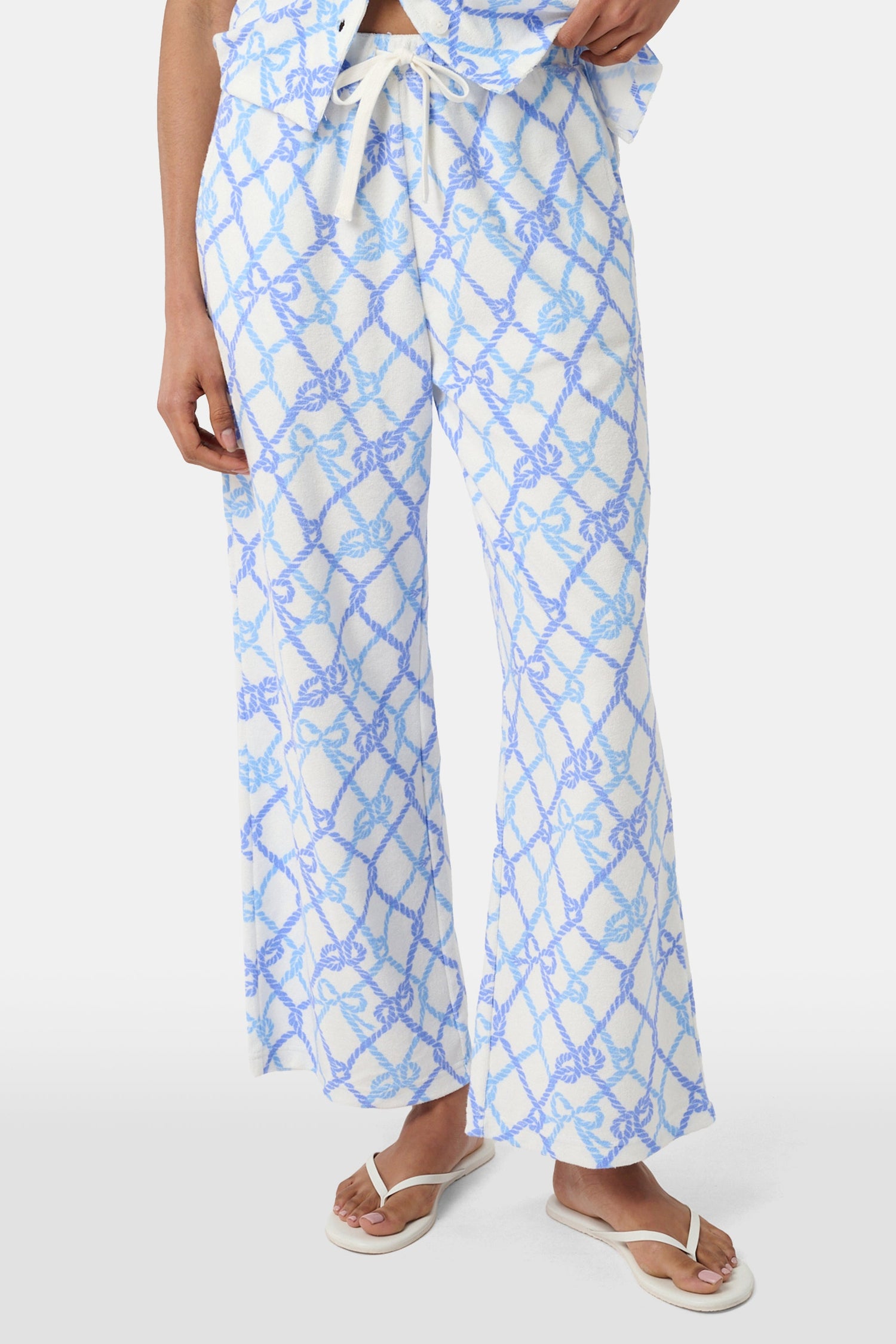 A person wears the Roller Rabbit Seaside Knots Mara Pant, featuring a tie waist and wide legs in loose white fabric with a light blue geometric chain pattern, paired with a matching top and white sandals against a plain white background.