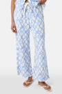 A person wears the Roller Rabbit Seaside Knots Mara Pant, featuring a tie waist and wide legs in loose white fabric with a light blue geometric chain pattern, paired with a matching top and white sandals against a plain white background.