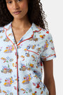 A woman with curly hair smiles in Roller Rabbit Spain Monkey Polo Pajamas—light blue shortie PJs made from 100 Pima cotton with colorful whimsical prints and red piping—standing against a plain white background.