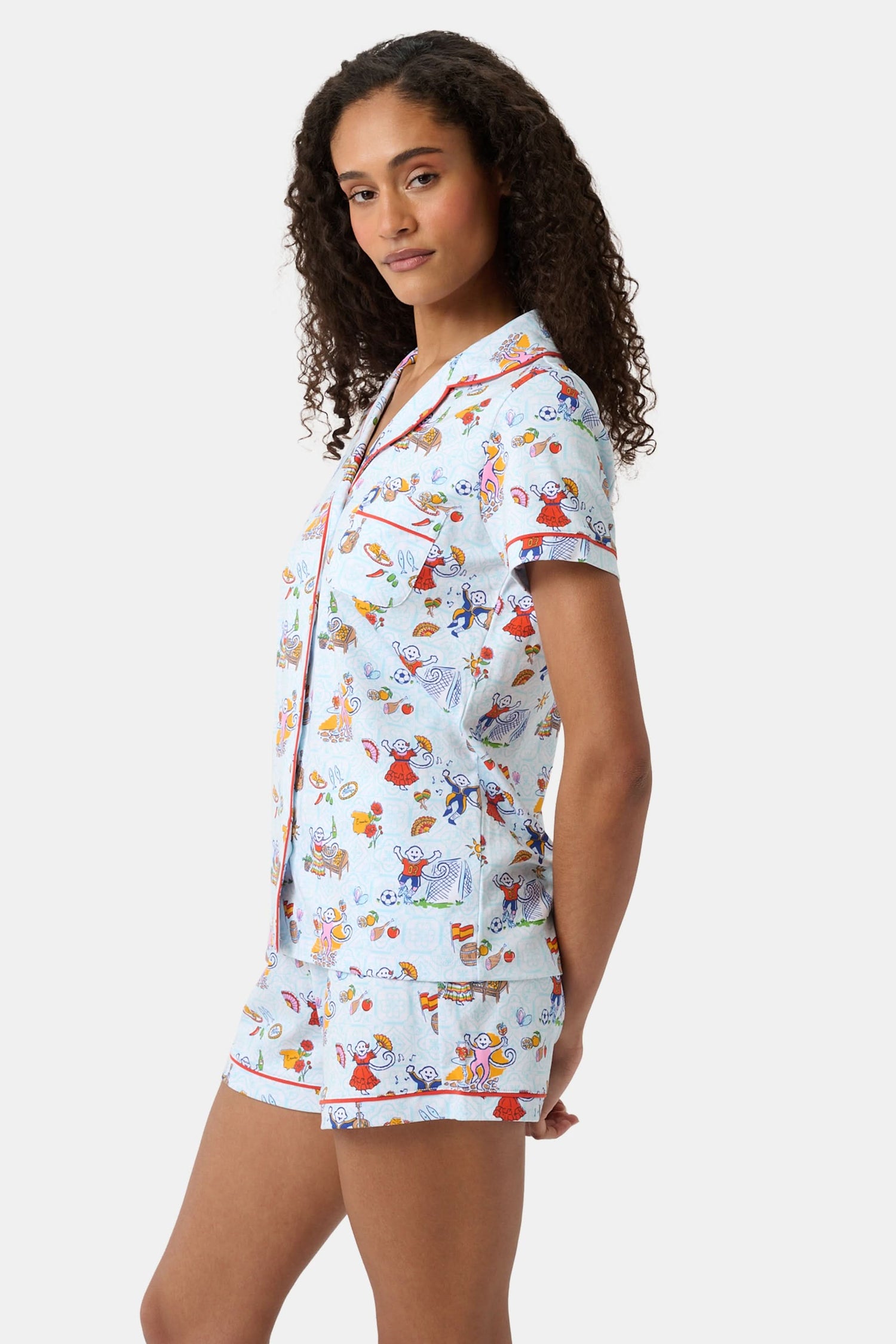 A woman with curly hair smiles in Roller Rabbit Spain Monkey Polo Pajamas—light blue shortie PJs made from 100 Pima cotton with colorful whimsical prints and red piping—standing against a plain white background.