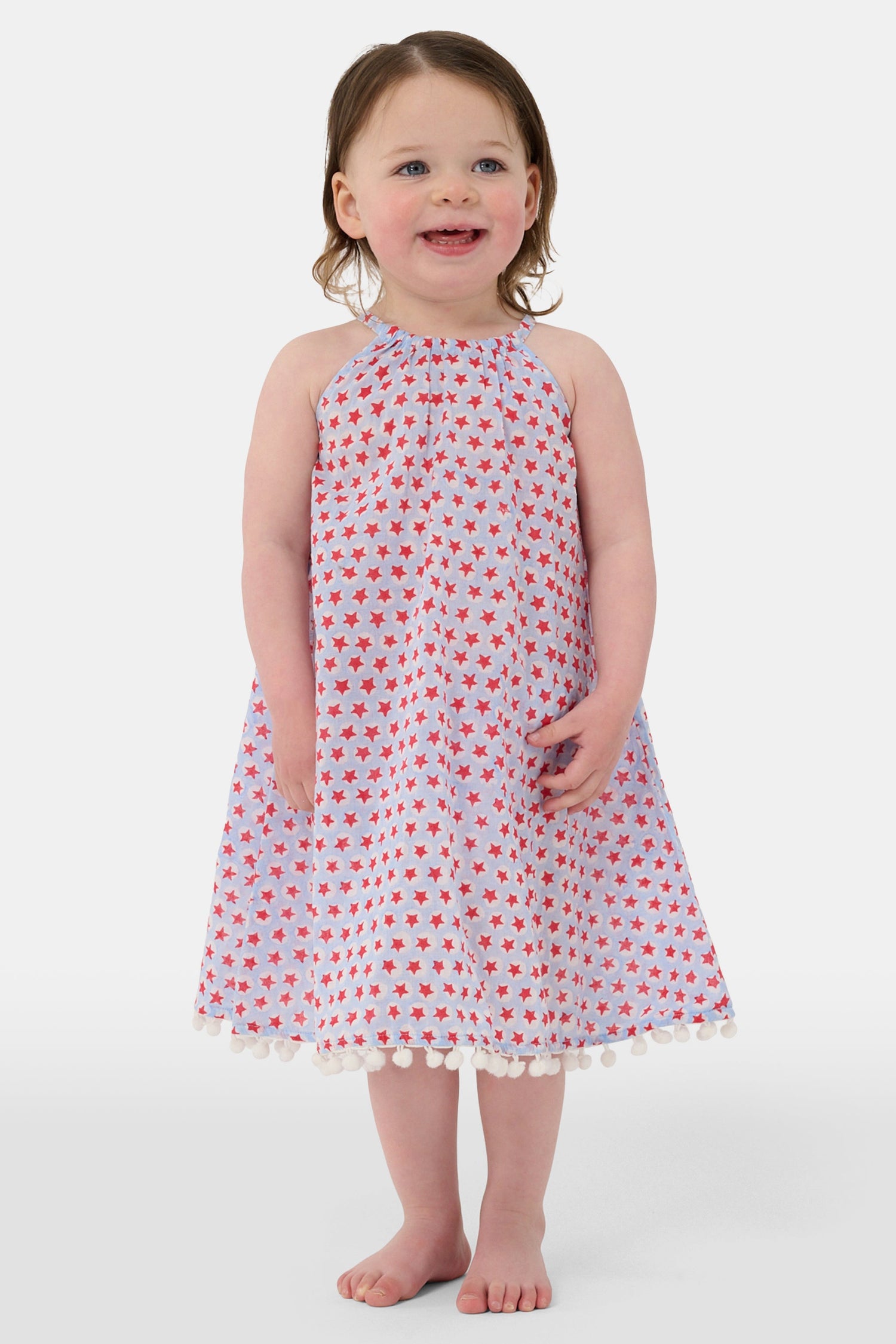 A young girl with long brown hair stands facing forward in the Roller Rabbit Kids Starry Dot Carrie Cover Up—a sleeveless, knee-length white dress featuring a red dotted pattern and pom-pom trim. She has a neutral expression and a light background behind her.