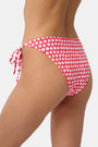 A person wears the Roller Rabbit Starry Dot Isa Bikini Bottom in red with white polka dots and adjustable hip ties, shown from mid-torso to upper thigh.
