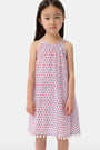 A young girl with long brown hair stands facing forward in the Roller Rabbit Kids Starry Dot Carrie Cover Up—a sleeveless, knee-length white dress featuring a red dotted pattern and pom-pom trim. She has a neutral expression and a light background behind her.
