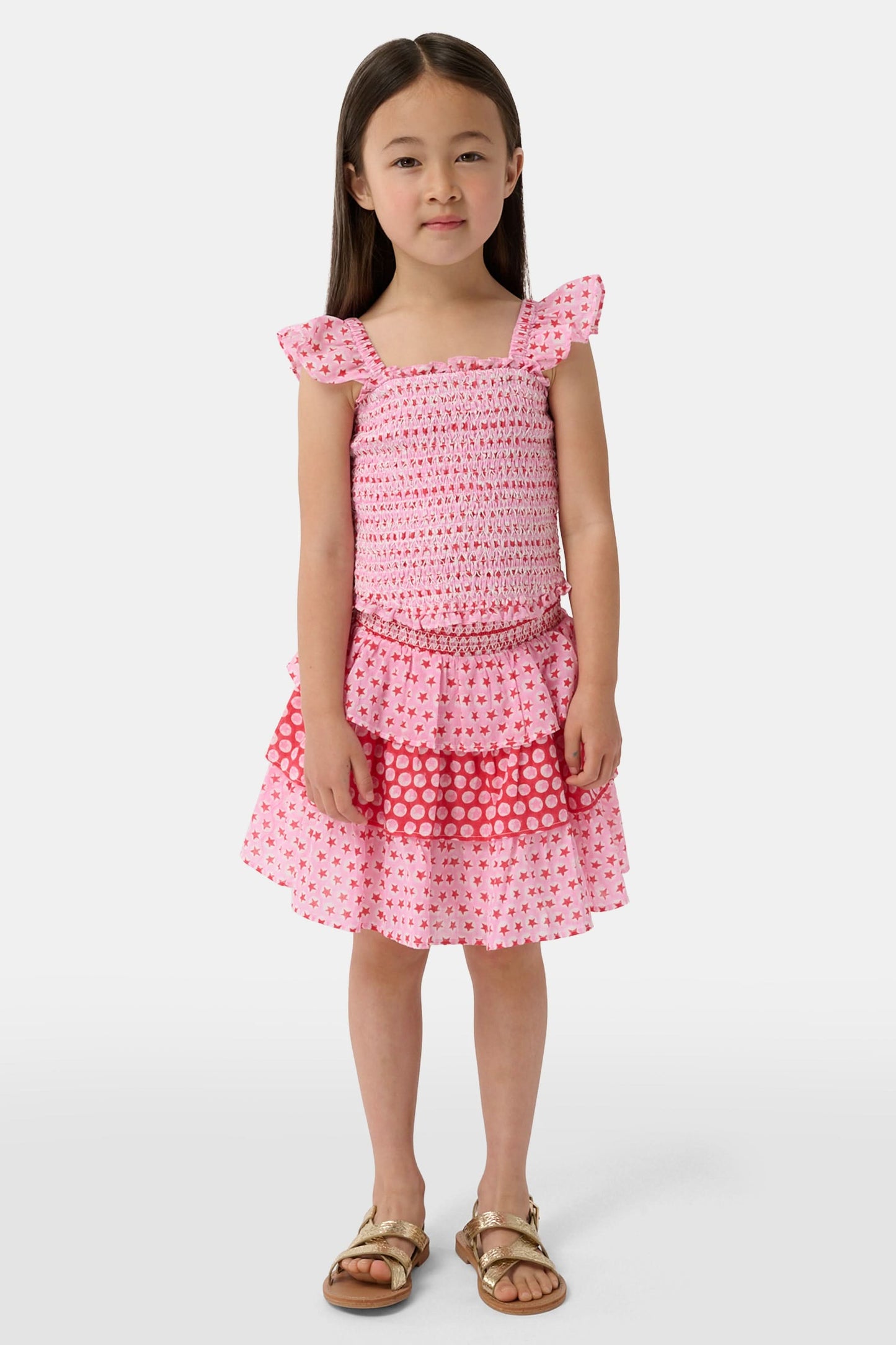 A child wears the Roller Rabbit Kids Starry Dot Sami Skirt in pink and red with star and polka dot patterns, paired with a pink smocked top, standing against a plain light background.