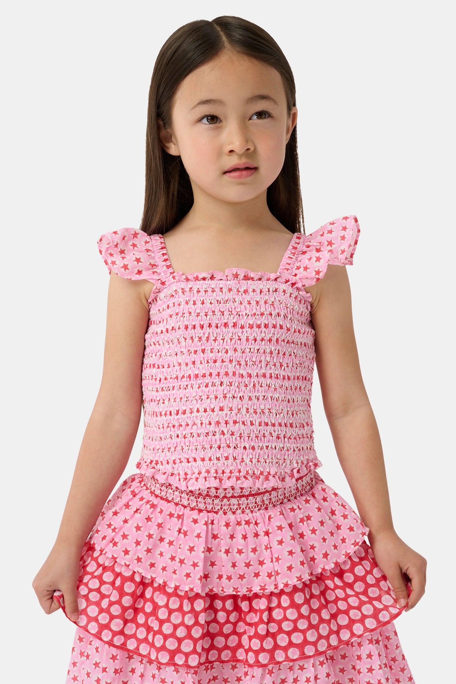 A young girl wears the Roller Rabbit Kids Starry Dot Tory Top in pastel pink with flutter sleeves, paired with a matching skirt featuring red stars and white polka dots. Only her torso and part of her face are visible.