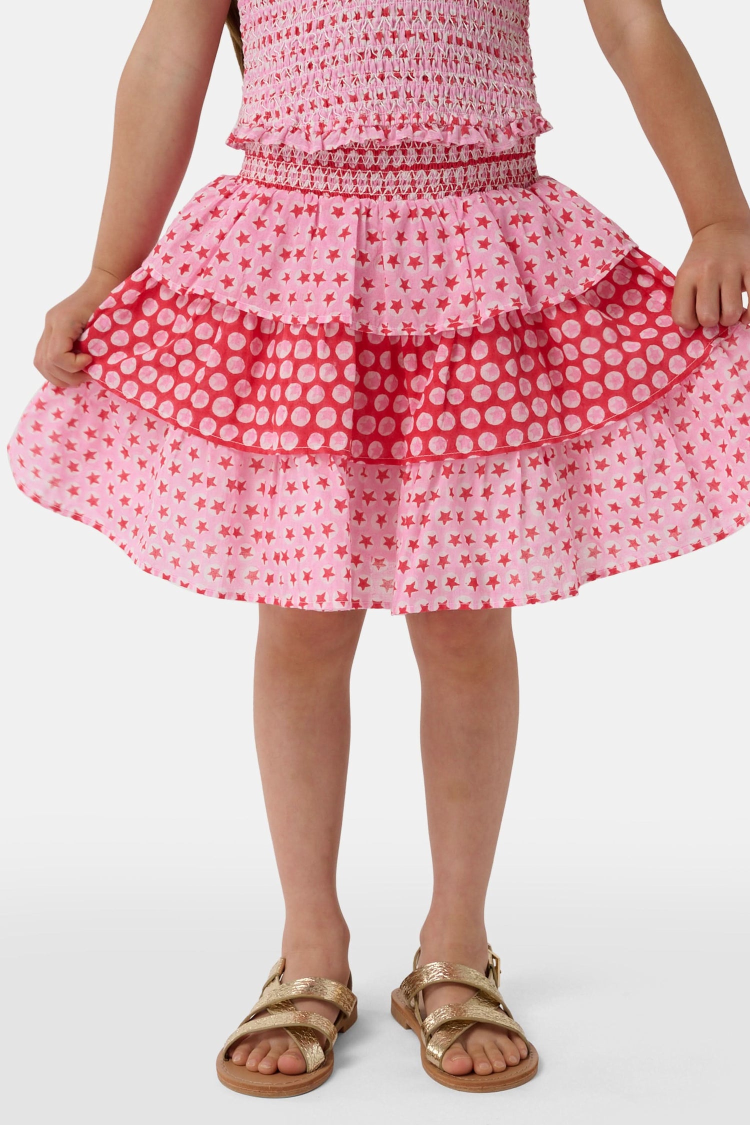 A child wears the Roller Rabbit Kids Starry Dot Sami Skirt in pink and red with star and polka dot patterns, paired with a pink smocked top, standing against a plain light background.