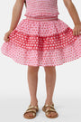 A child wears the Roller Rabbit Kids Starry Dot Sami Skirt in pink and red with star and polka dot patterns, paired with a pink smocked top, standing against a plain light background.