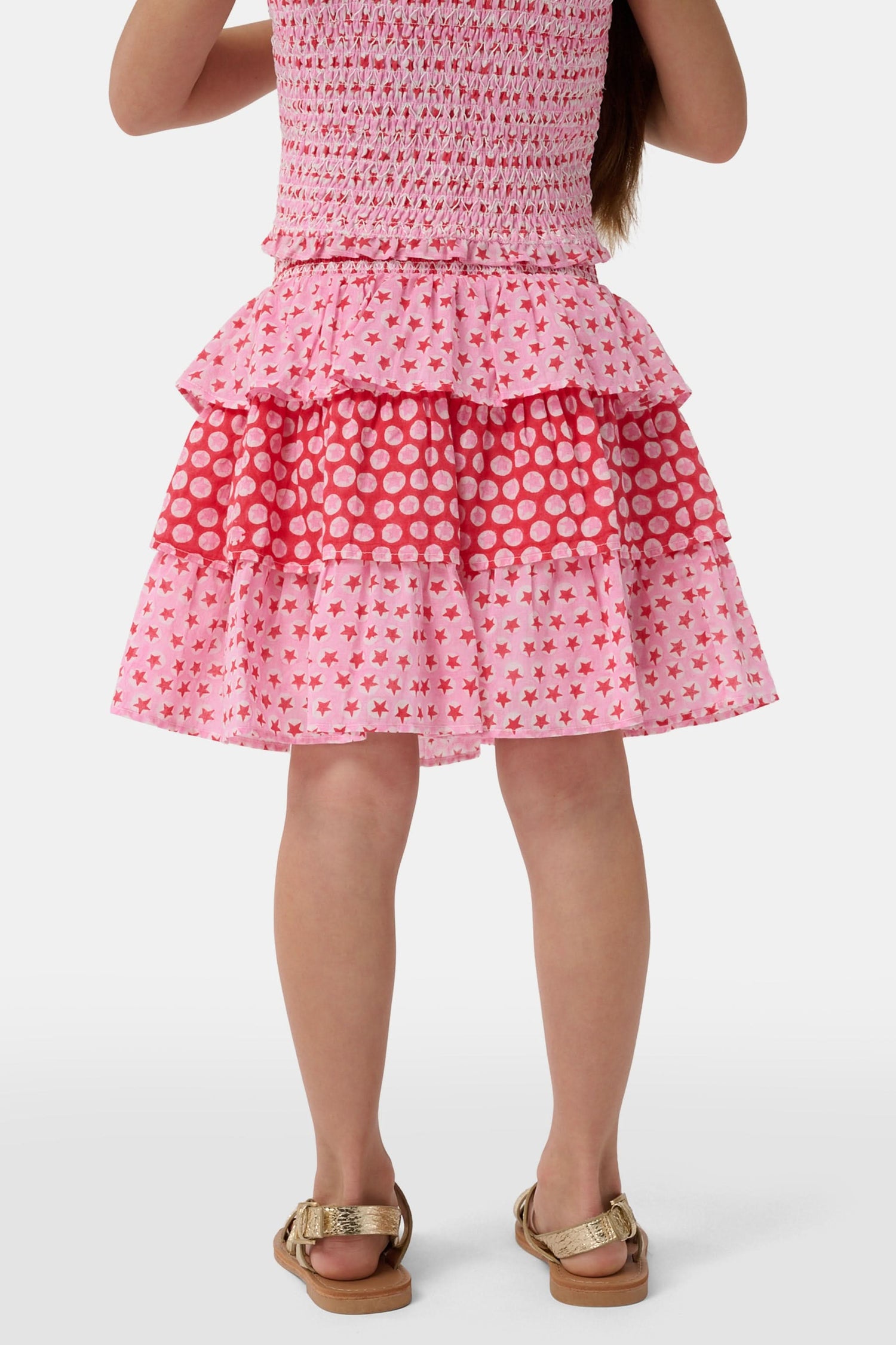 A child wears the Roller Rabbit Kids Starry Dot Sami Skirt in pink and red with star and polka dot patterns, paired with a pink smocked top, standing against a plain light background.