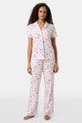 A woman with curly hair models the Roller Rabbit Summer Memories Fiora Pajamas, a Pima cotton set with a colorful sushi print, including a short-sleeve button-up top and long pants, against a white background.