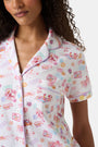 A woman with curly hair models the Roller Rabbit Summer Memories Fiora Pajamas, a Pima cotton set with a colorful sushi print, including a short-sleeve button-up top and long pants, against a white background.