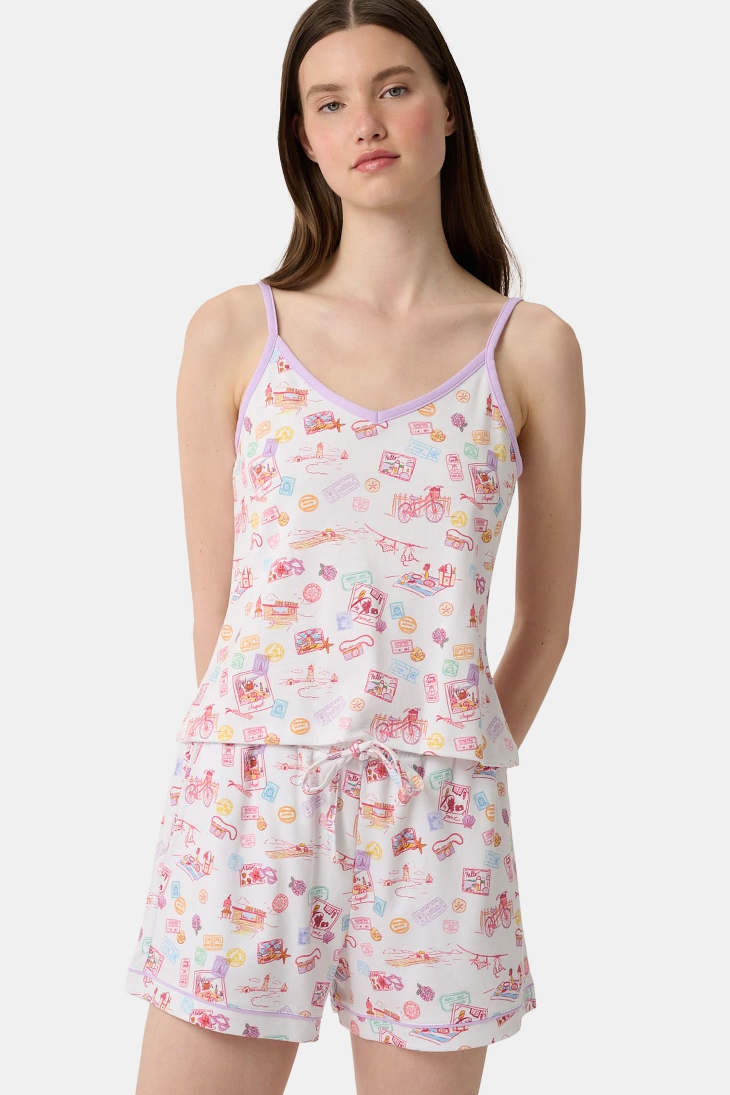 A woman wears the Roller Rabbit Summer Memories Mischa Set, a 100 Pima cotton cami tank and boxer set with a pastel travel icon print on white, standing against a light background.