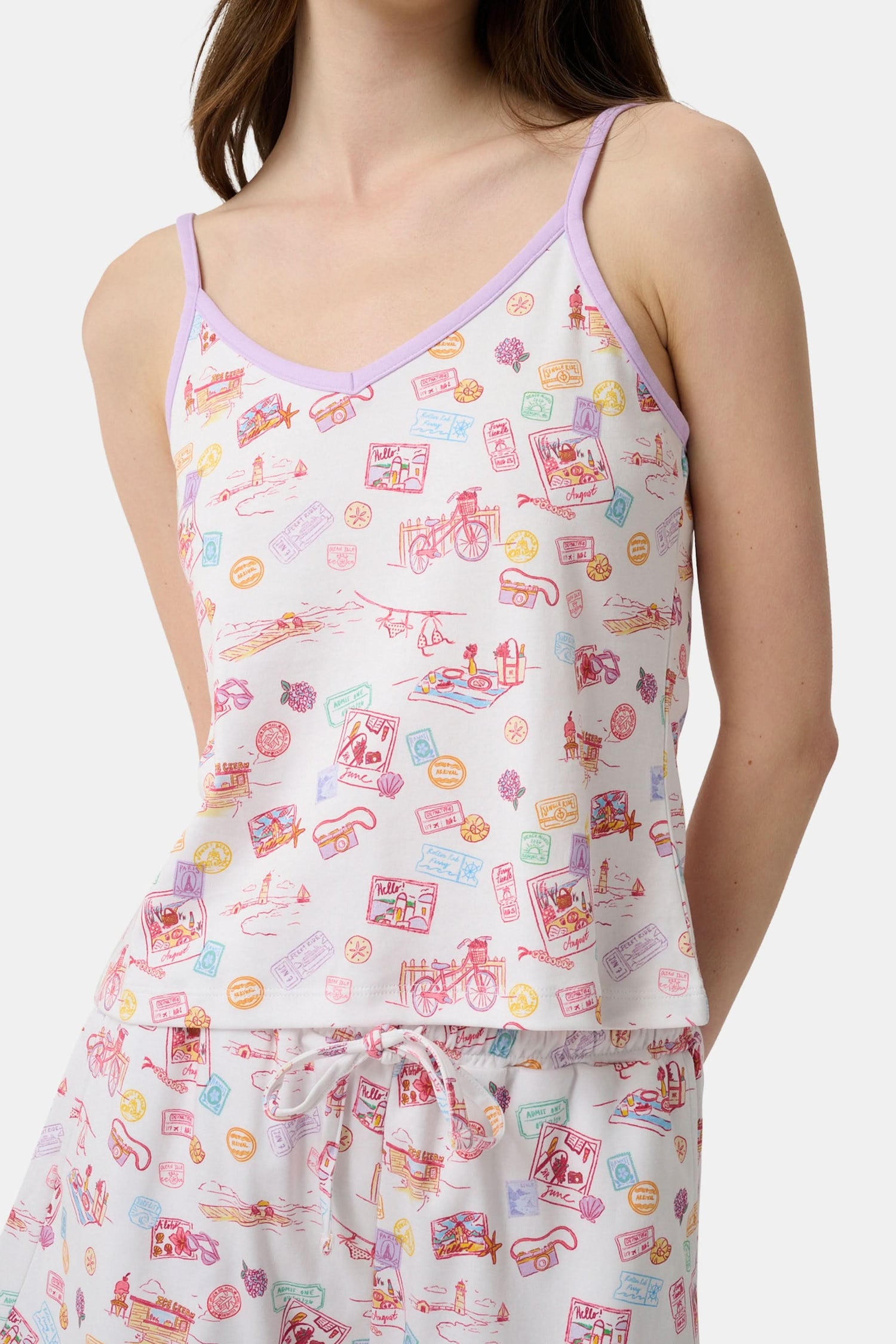 A woman wears the Roller Rabbit Summer Memories Mischa Set, a 100 Pima cotton cami tank and boxer set with a pastel travel icon print on white, standing against a light background.