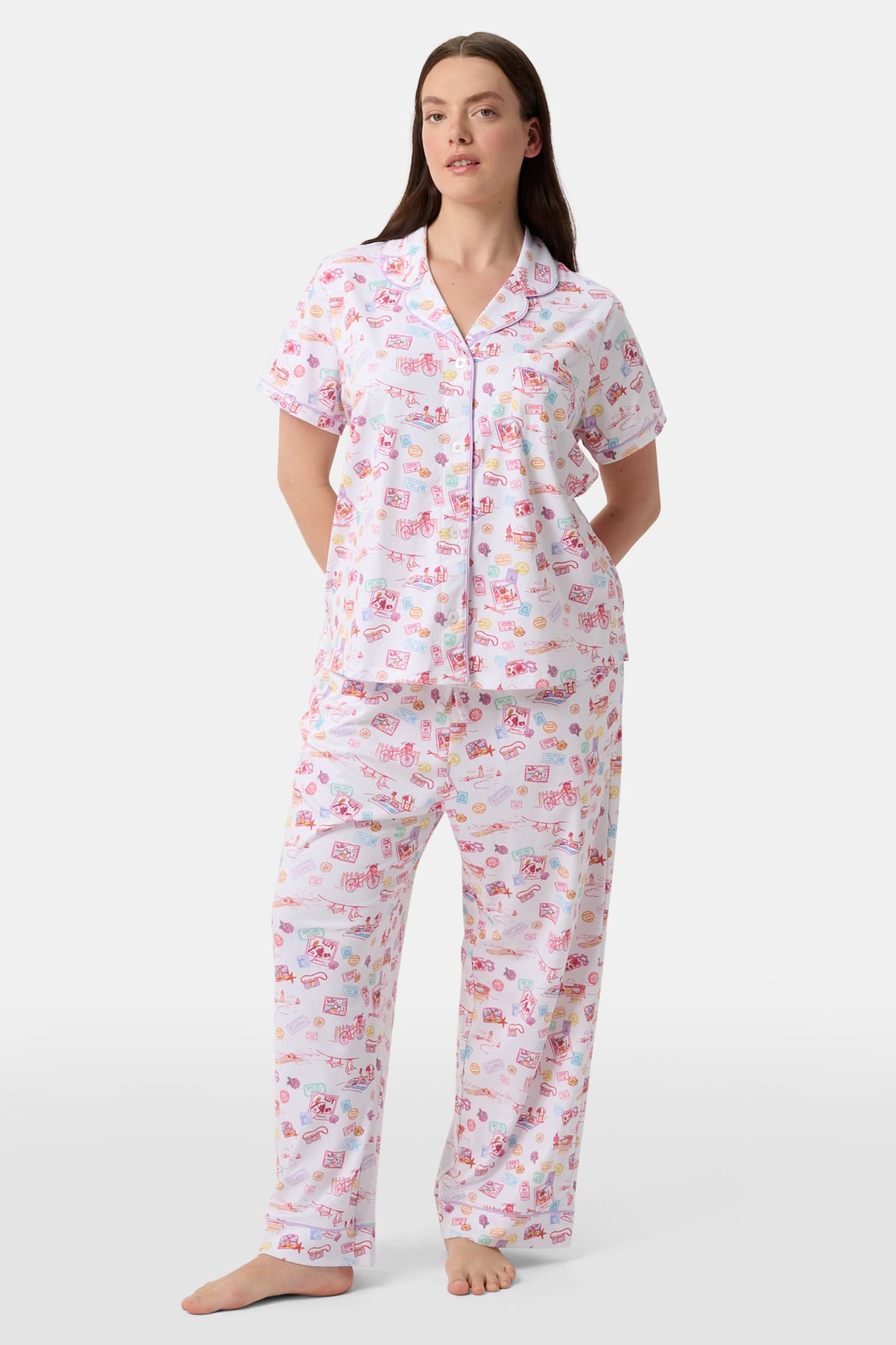 A woman with curly hair models the Roller Rabbit Summer Memories Fiora Pajamas, a Pima cotton set with a colorful sushi print, including a short-sleeve button-up top and long pants, against a white background.