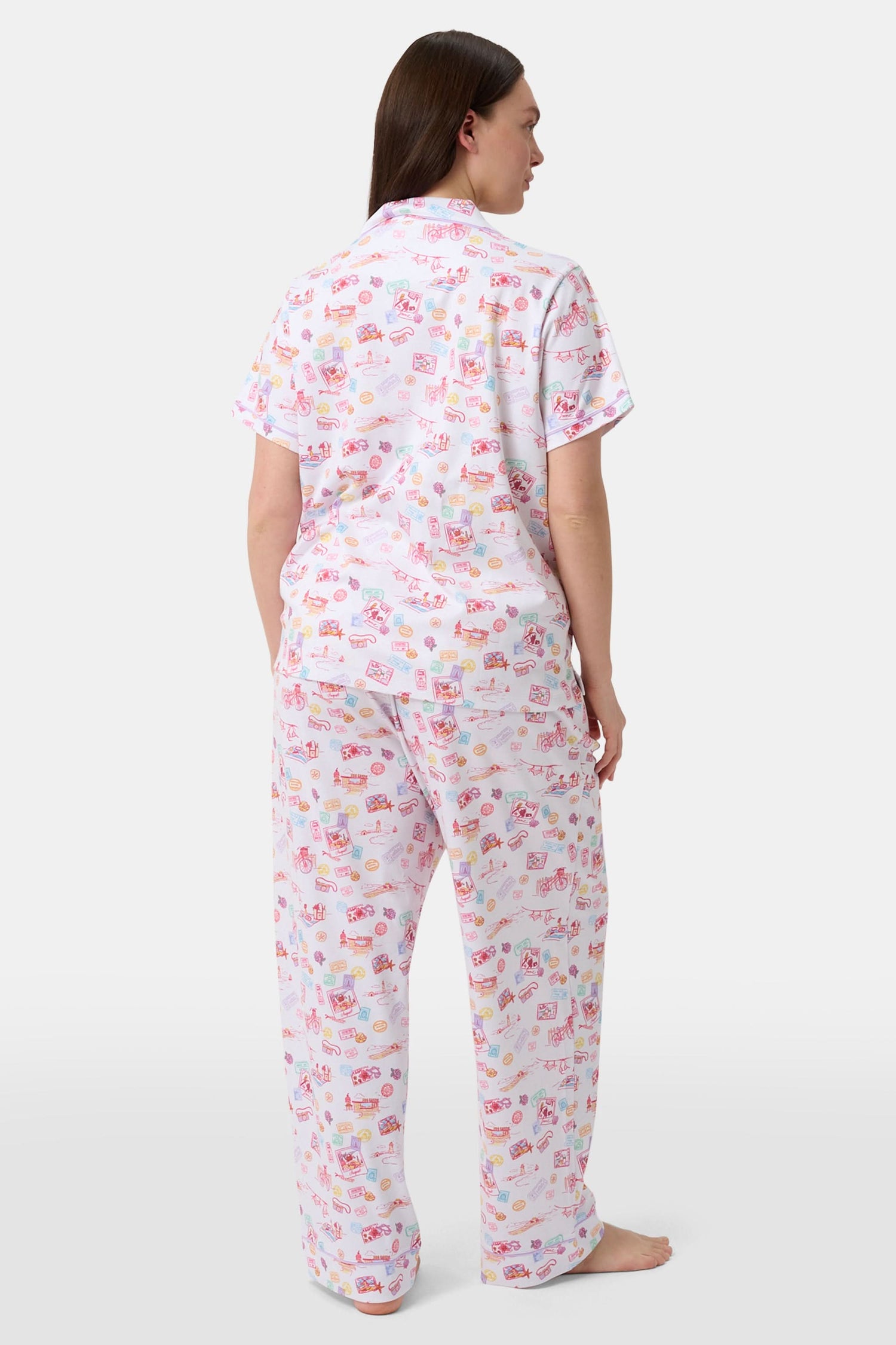 A woman with curly hair models the Roller Rabbit Summer Memories Fiora Pajamas, a Pima cotton set with a colorful sushi print, including a short-sleeve button-up top and long pants, against a white background.