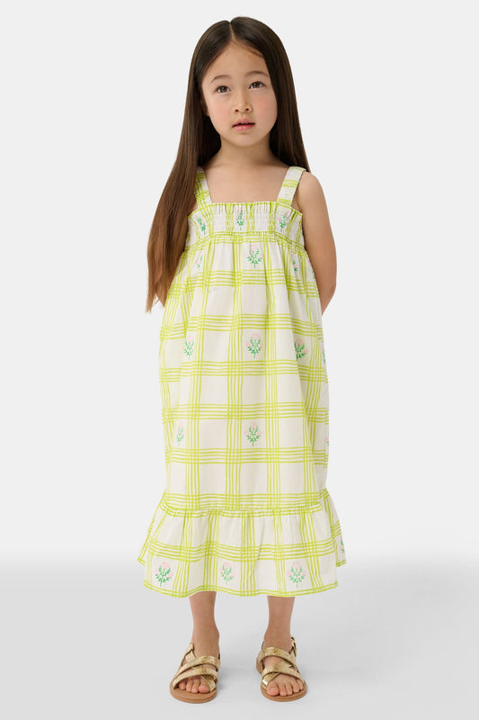 A young girl with long brown hair wears the Roller Rabbit Kids Sun Check Blossom Teagan Dress—a yellow-and-white plaid sundress with ruffled hem and green floral accents. She stands facing forward, hands behind her back, in gold sandals.