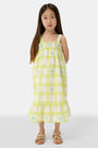 A young girl with long brown hair wears the Roller Rabbit Kids Sun Check Blossom Teagan Dress—a yellow-and-white plaid sundress with ruffled hem and green floral accents. She stands facing forward, hands behind her back, in gold sandals.