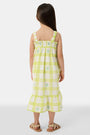 A young girl with long brown hair wears the Roller Rabbit Kids Sun Check Blossom Teagan Dress—a yellow-and-white plaid sundress with ruffled hem and green floral accents. She stands facing forward, hands behind her back, in gold sandals.